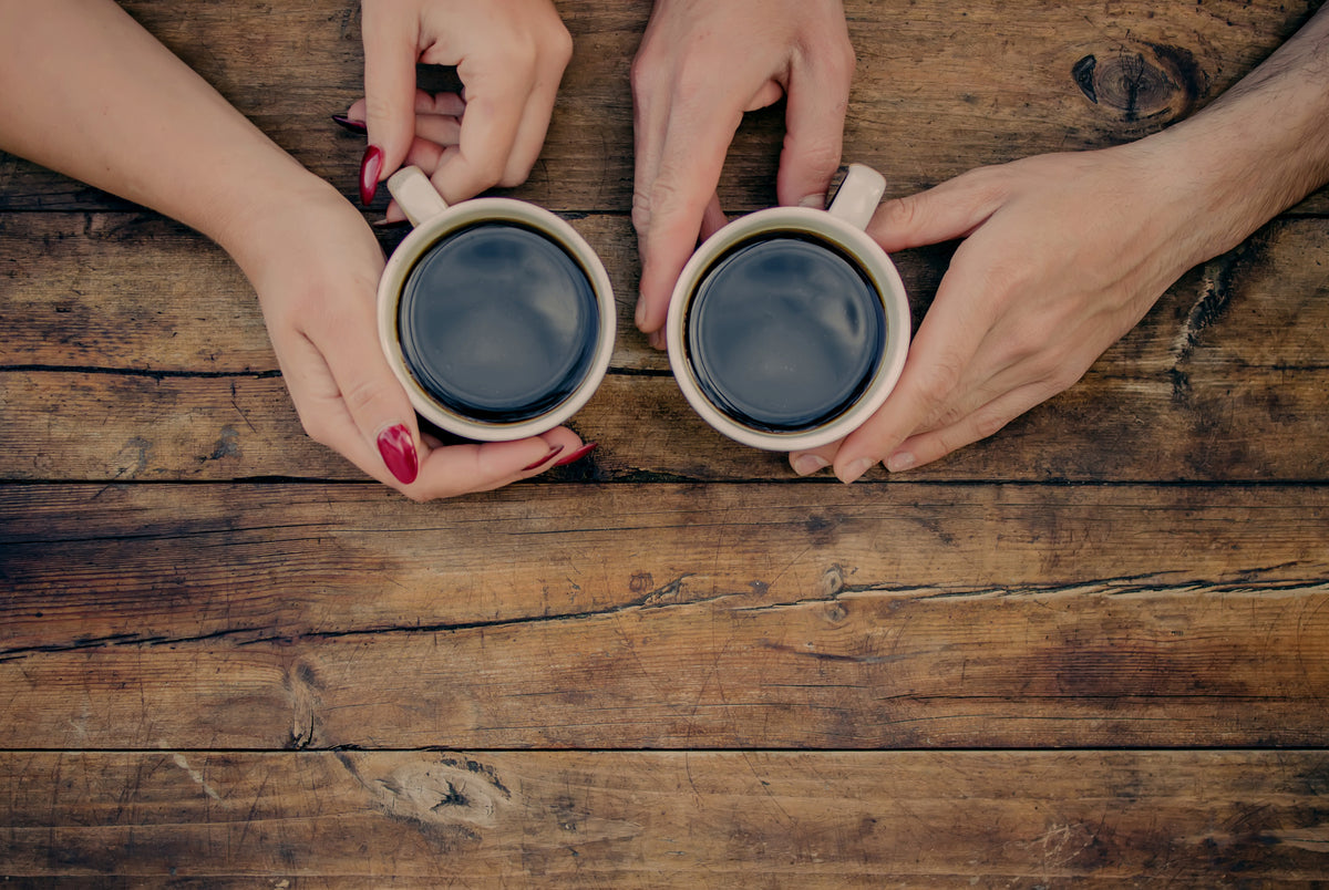 Two people holding cups of black coffee at a wooden table